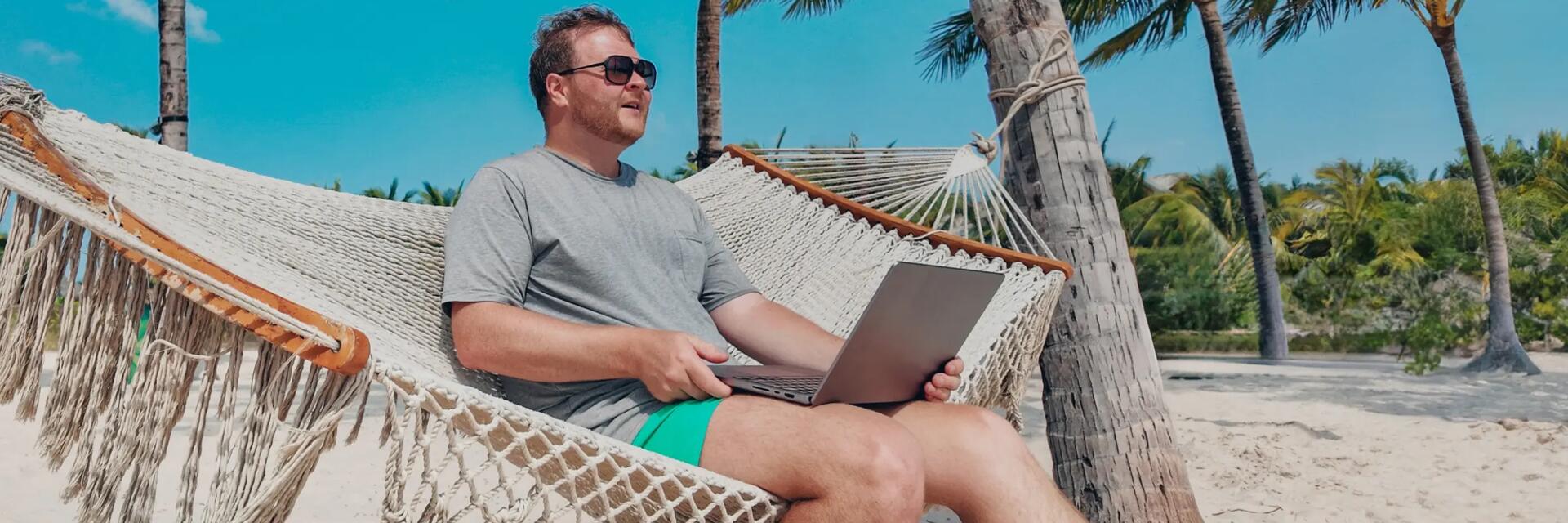 Mężczyzna siedzący na hamaku wśród palm z laptopem na kolanach