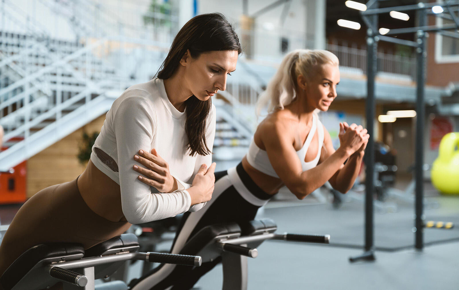 Dwie kobiety w odzieży sportowej ćwiczące unoszenie tułowia na ławce rzymskiej na siłowni