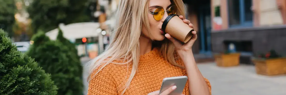 Kobieta pijąca z kuba jednorazowego w drugiej ręce trzymająca telefon