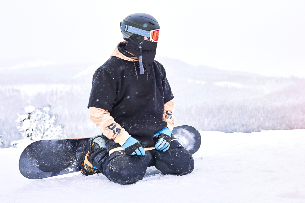 Mężczyzna w kasku i w kominiarce siedzący na kolanach z deską snowboardową przypiętą do stóp na śniegu