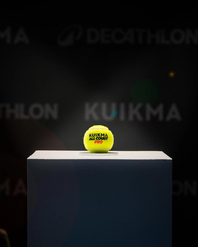 Ein gelber Tennisball liegt mittig auf einem weißen Sockel vor einem dunklen Hintergrund. Auf dem Ball steht "Kuikma All Court Pro".
