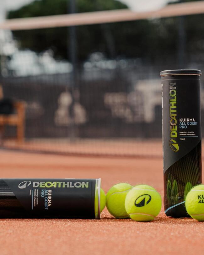 Zwei schwarze Dosen und gelbe Tennisbälle liegen auf einem rötlichen Sandplatz vor einem Tennisnetz. Auf der Verpackung und den Bällen ist das Logo sowie der Markenname „Decathlon“ erkennbar. Ein Tennisball im Vordergrund trägt die spezifische Aufschrift „KUIKMA ALL COURT PRO“.