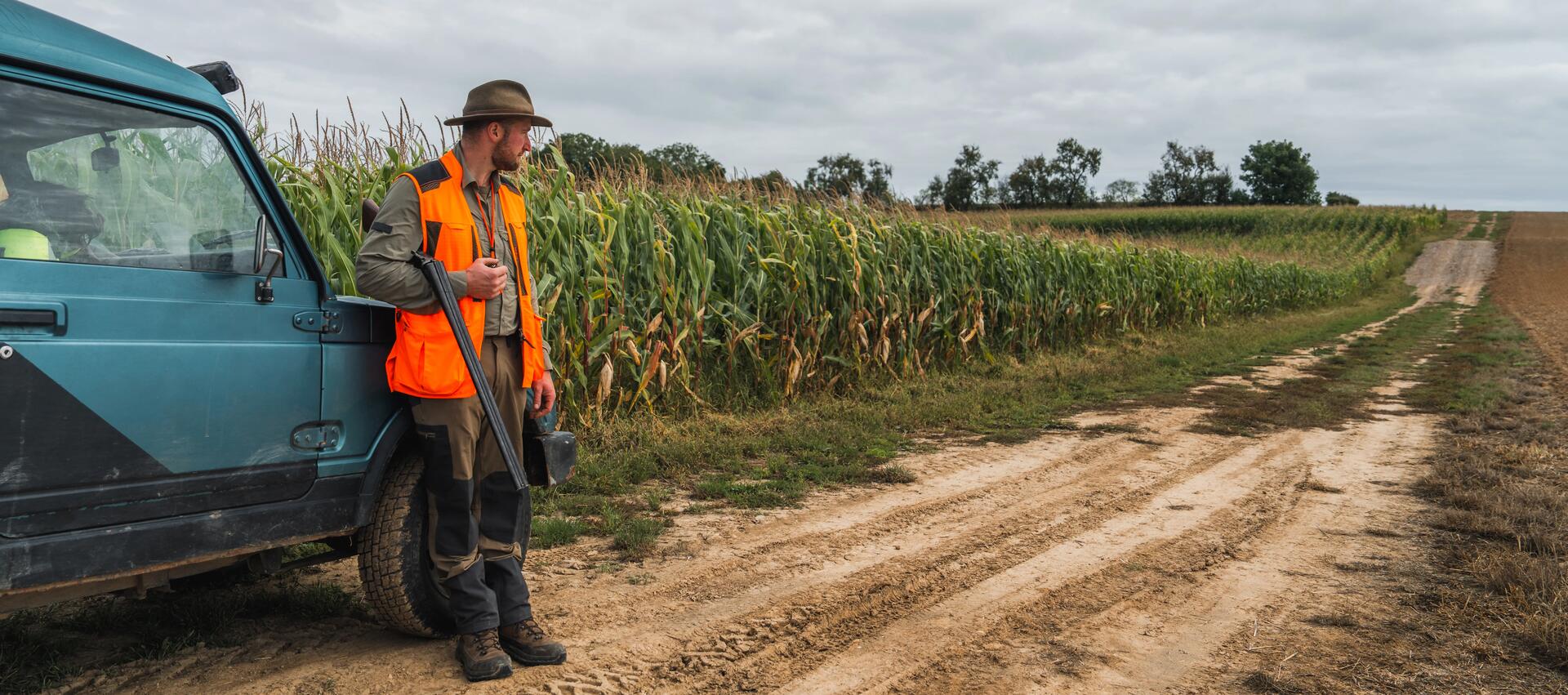 un chasseur avec son gilet orange fluo sur le dos près d'un champ de maïs et adossé à une voiture