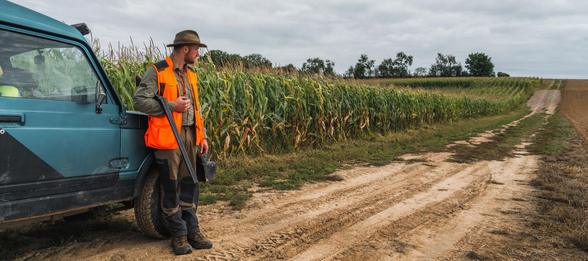 un chasseur avec son gilet orange fluo sur le dos près d'un champ de maïs et adossé à une voiture