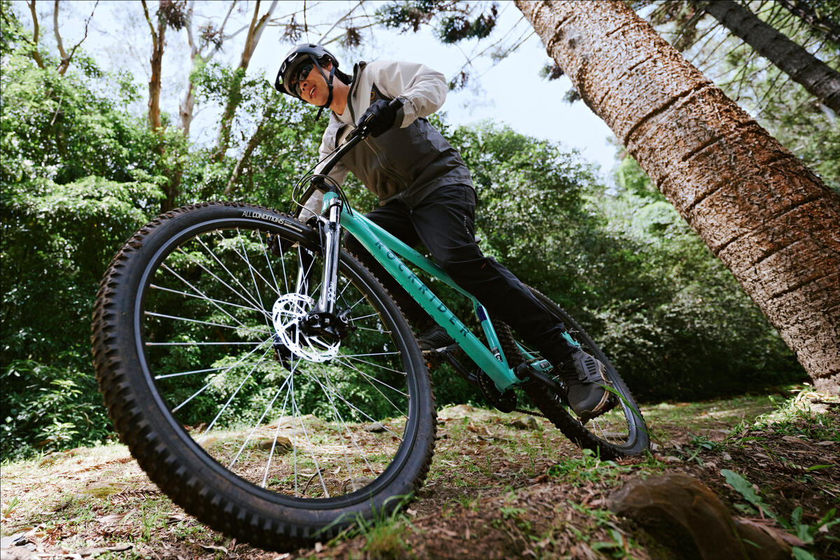 Oseba s čelado in športno opremo vozi gorsko kolo po zemeljski poti skozi s soncem obsijano gozd na turkiznem kolesu Rockrider.