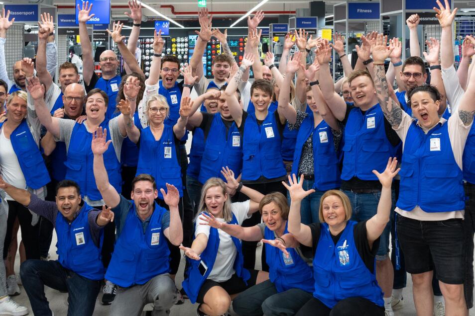 Eine große, vielfältige Gruppe von lächelnden Mitarbeitern in einem Sportgeschäft. Sie tragen alle einheitliche blaue Westen über ihrer Alltagskleidung. Fast alle Personen haben die Arme jubelnd in die Luft gestreckt oder winken freudig in die Kamera. Die Gruppe ist in mehreren Reihen gestaffelt, wobei einige im Vordergrund knien. Im Hintergrund sind Verkaufsregale und Abteilungsschilder wie „Wassersport“, „Fitness“ und „Bekleidung“ zu sehen. Die Atmosphäre ist energiegeladen und teamorientiert.