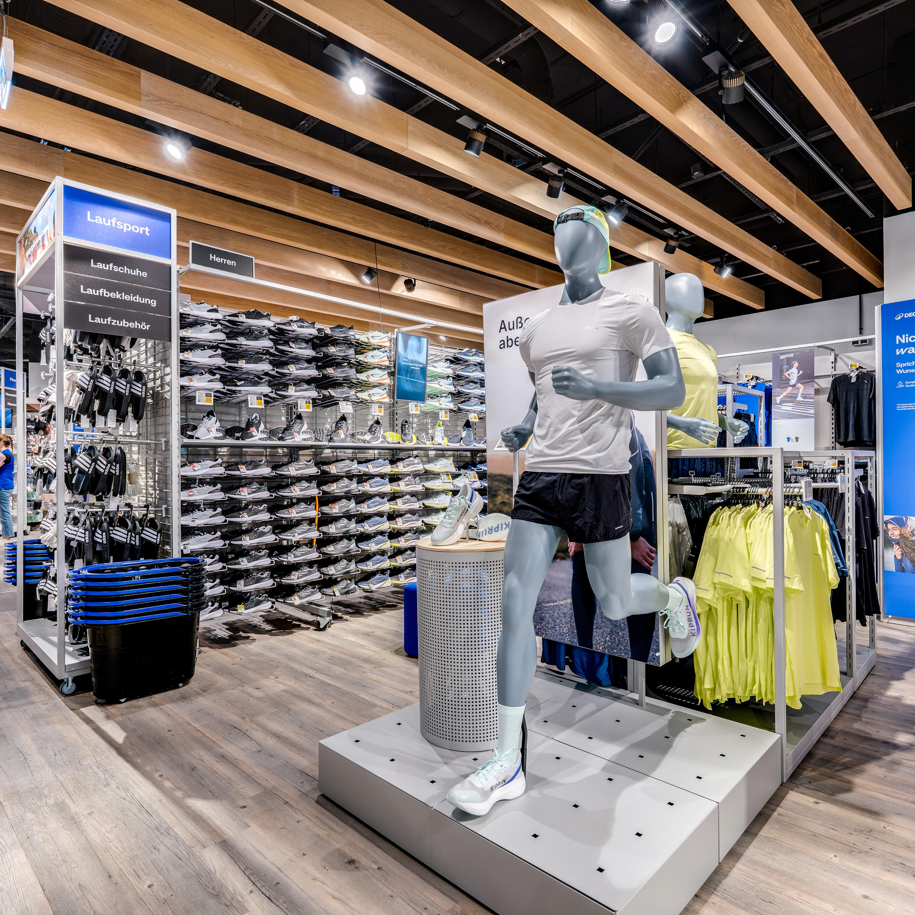 Blick in die moderne Laufsport-Abteilung eines Sportgeschäfts. Im Vordergrund steht eine männliche Schaufensterpuppe in dynamischer Laufpose, bekleidet mit weißem T-Shirt, schwarzen Shorts und Laufschuhen. Dahinter befinden sich gut sortierte Regale mit einer großen Auswahl an Laufschuhen für Herren sowie Ständer mit neongelber Sportbekleidung. Die Decke ist mit hellen Holzpaneelen und moderner Spot-Beleuchtung gestaltet, was dem Verkaufsraum eine offene und freundliche Atmosphäre verleiht.