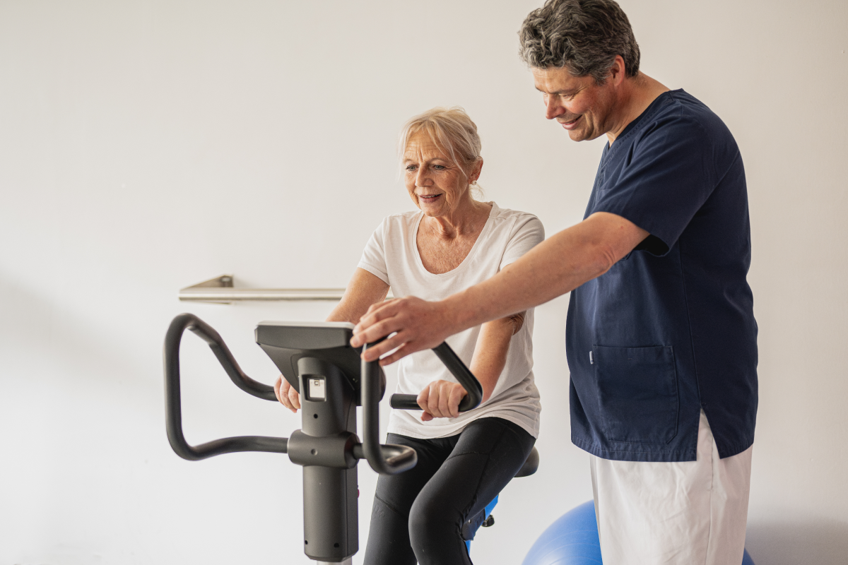 Kobieta ćwicząca na rowerze stacjonarnym obok rehabilitant