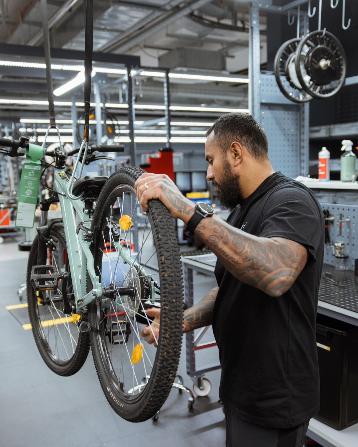 Ein Mechaniker mit tätowierten Armen und einer Smartwatch am Handgelenk arbeitet in einer hell erleuchteten Fahrradwerkstatt. Er hält das Hinterrad eines hellgrünen Mountainbikes fest, das an Gurten von der Decke hängt. Im Hintergrund sind Werkzeuge, Regale und weitere Fahrradteile zu sehen.