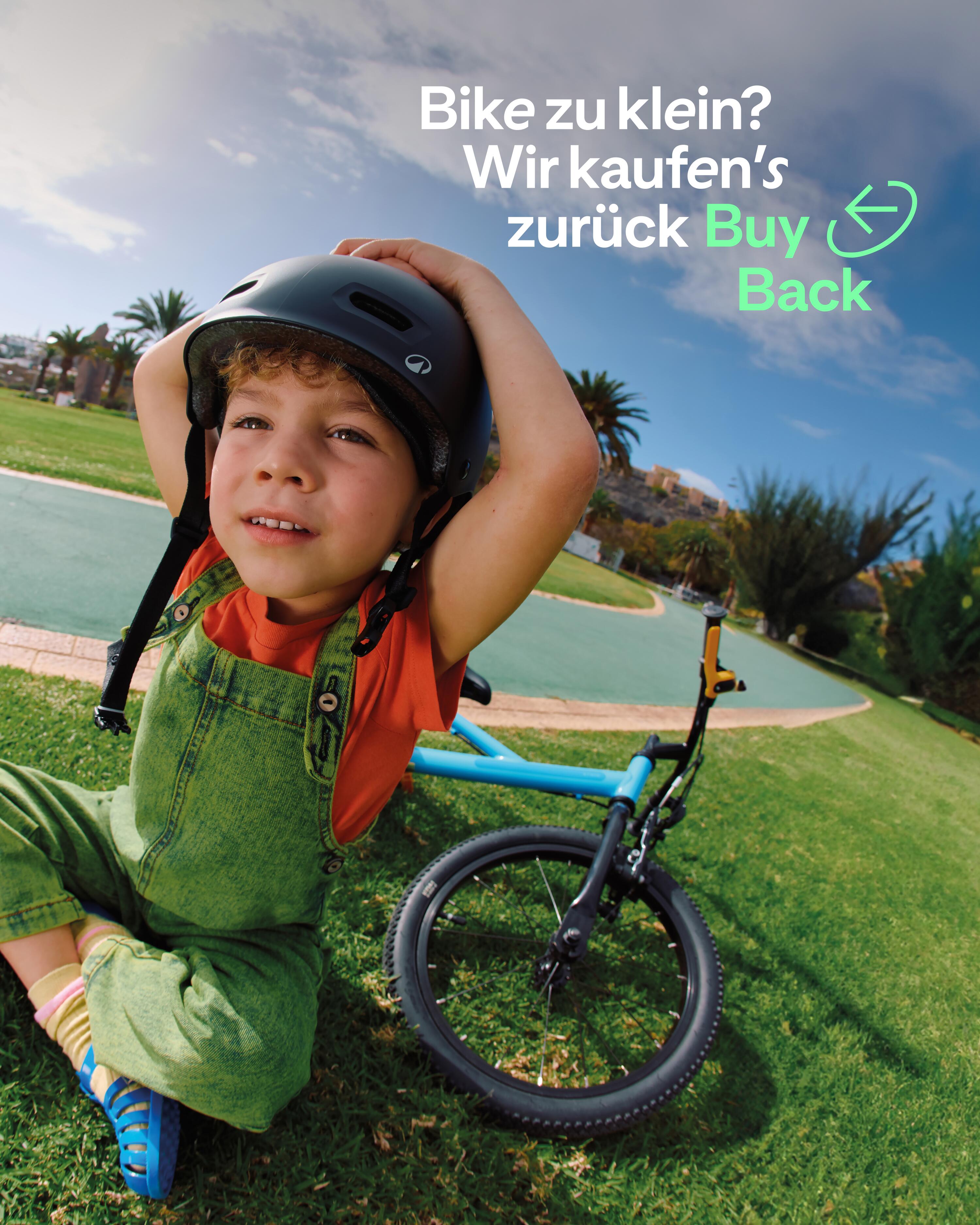 Ein Kind mit Locken und einem schwarzen Fahrradhelm sitzt lächelnd auf einer Rasenfläche neben einem kleinen blauen Fahrrad. Es trägt eine grüne Latzhose über einem orangefarbenen T-Shirt. Im Hintergrund ist ein sonniger Park mit Palmen und einem gepflasterten Weg zu sehen. Oben rechts steht der Text: „Bike zu klein? Wir kaufen’s zurück Buy Back“.