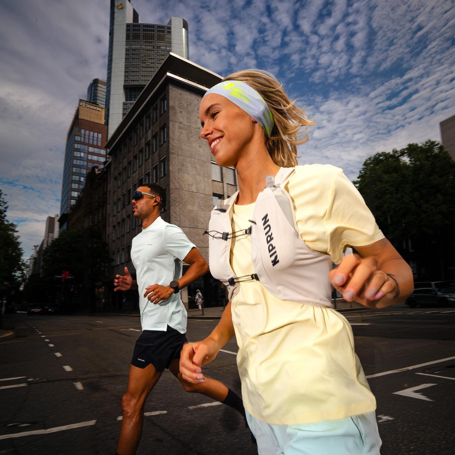 Zwei Personen joggen dynamisch durch eine urbane Umgebung mit hohen Gebäuden im Hintergrund. Im Fokus steht eine lächelnde Frau im Vordergrund, die ein gelbes T-Shirt, eine graue Laufweste der Marke Kiprun und ein Stirnband trägt. Hinter ihr läuft ein Mann in einem hellblauen Sportshirt. Die Kameraperspektive ist leicht von unten angesetzt, was zusammen mit dem bewölkten Himmel und der Architektur eine moderne, sportliche Großstadt-Atmosphäre erzeugt.