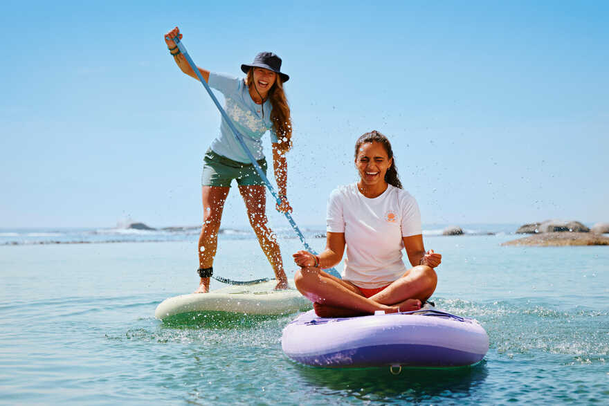 Dve nasmejane žene na SUP daskama; jedna stoji i vesla, a druga sedi.