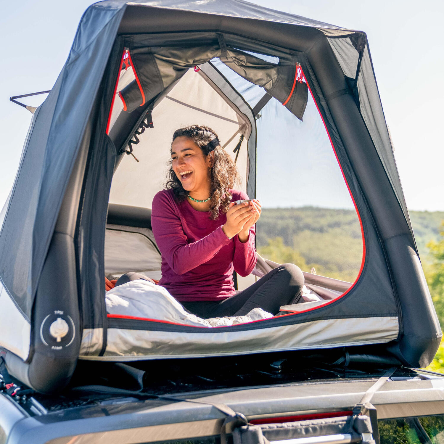 Eine lachende Frau sitzt in einem aufgeblasenen Dachzelt auf einem Autodach und hält eine Tasse in den Händen. Das moderne Zelt bietet einen weiten Ausblick auf eine grüne Hügellandschaft bei sonnigem Wetter. Im Vordergrund ist die geöffnete Zeltöffnung mit roten Akzenten und die Heckpartie des Fahrzeugs zu sehen.