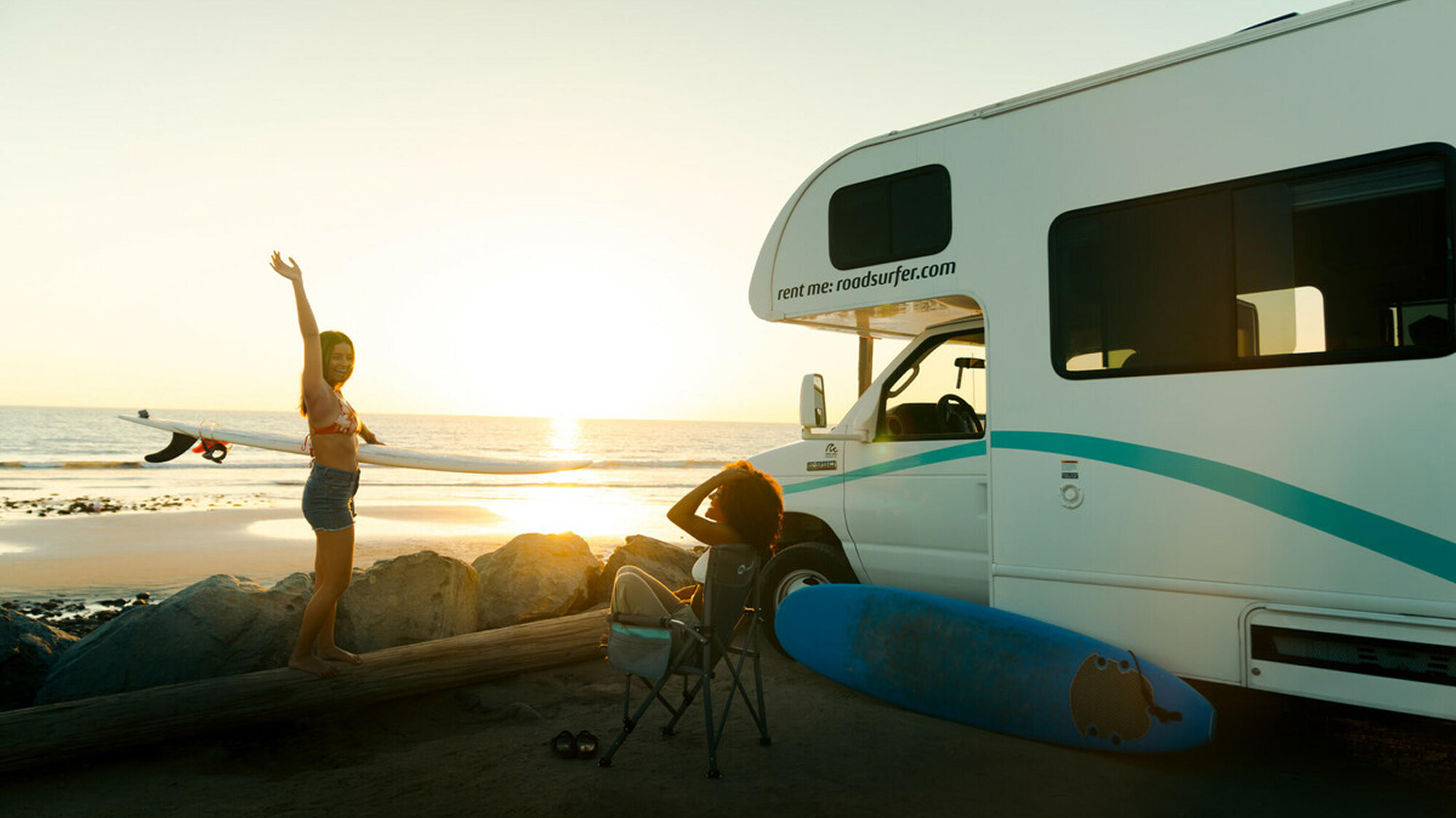Zwei Frauen entspannen am Strand mit einem roadsurfer-Wohnmobil bei Sonnenuntergang. Sie halten Surfbretter und genießen die Aussicht auf das Meer.