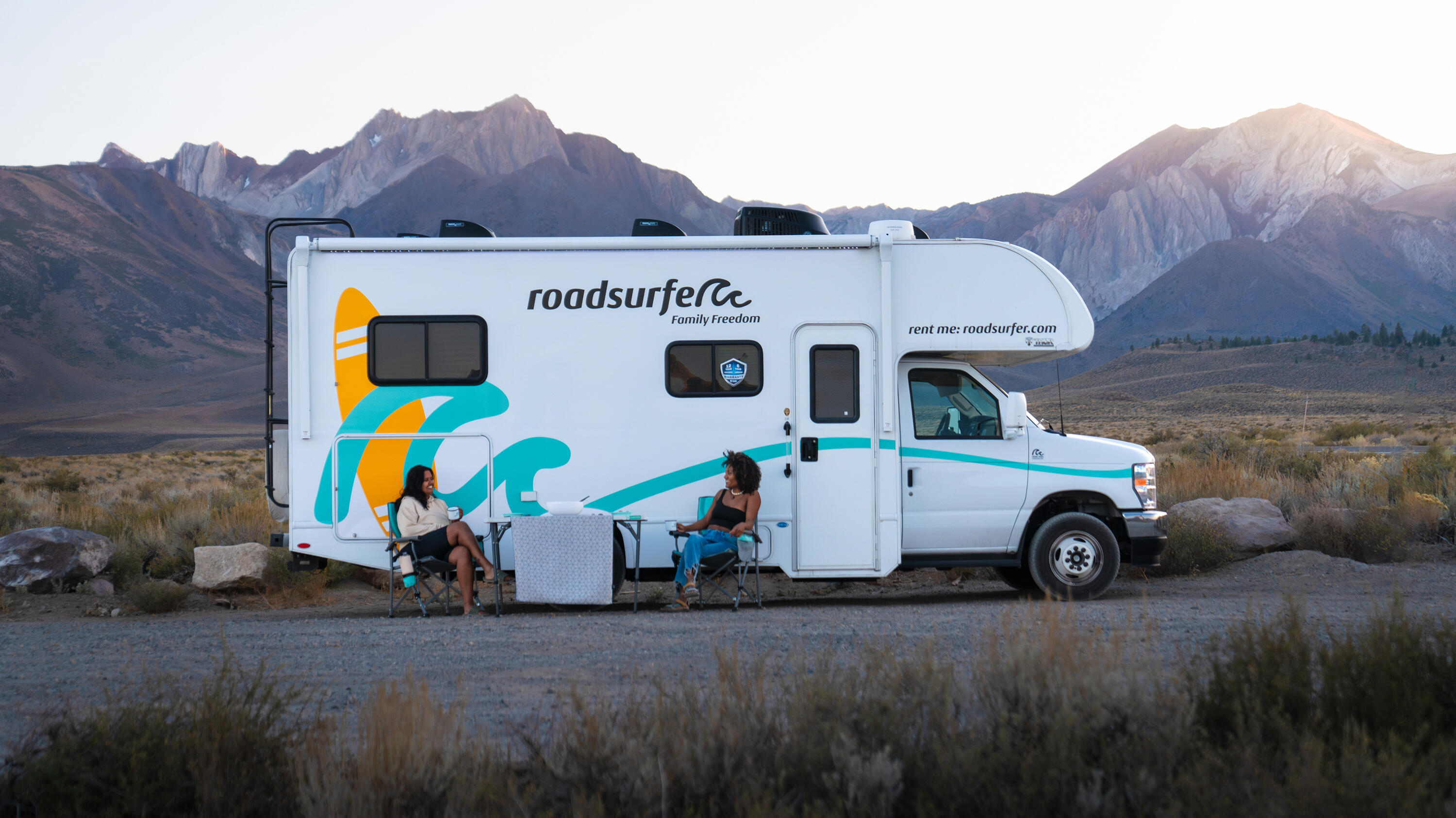 Roadsurfer-Wohnmobil mit Camping-Set vor einer spektakulären Bergkulisse bei Sonnenuntergang.