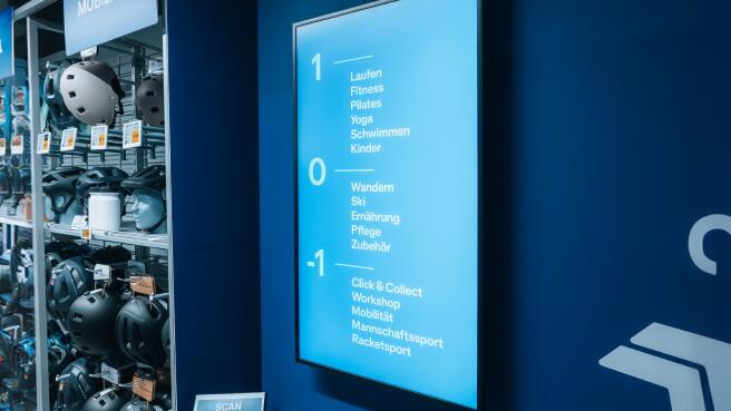 Innenansicht eines Sportgeschäfts mit Fokus auf eine digitale Etagenübersicht an einer blauen Wand. Der Bildschirm listet Abteilungen für drei Stockwerke auf:  Ebene 1: Laufen, Fitness, Pilates, Yoga, Schwimmen, Kinder.  Ebene 0: Wandern, Ski, Ernährung, Pflege, Zubehör.  Ebene -1: Click & Collect, Workshop, Mobilität, Mannschaftssport, Racketsport.