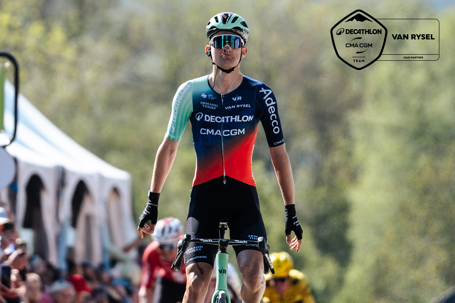 Ein professioneller Radrennfahrer im blau-rot-weißen Trikot von „Decathlon AG2R La Mondiale“ überquert jubelnd mit erhobenem Arm die Ziellinie, während im Hintergrund weitere Fahrer folgen und Zuschauer hinter blauen Werbebanden am Straßenrand stehen. Die Szene zeigt ein Straßenradrennen bei Tageslicht mit Fokus auf den Sieger in der Bildmitte.