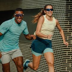 Zwei gut gelaunte Personen beim Lauftraining vor einer gefliesten Wand. Eine Frau im Vordergrund und ein Mann dahinter laufen dynamisch und lachen. Beide tragen sportliche Kleidung – die Frau ein hellgelbes Tanktop und dunkle Laufshorts, der Mann ein türkisfarbenes T-Shirt und helle Shorts – sowie dunkle Sportsonnenbrillen.