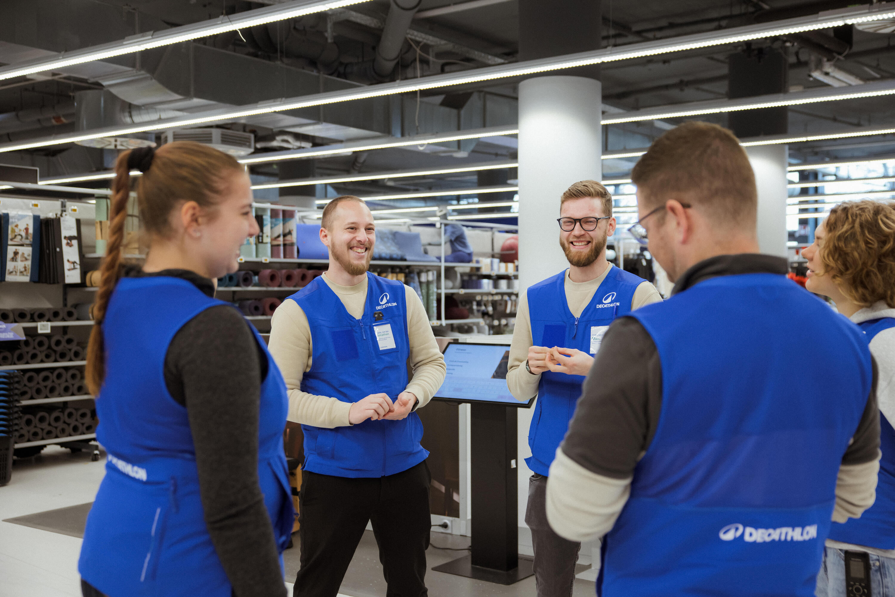 Eine Gruppe von fünf Decathlon-Mitarbeitenden in blauen Firmenwesten steht in einem Kreis in der Filiale und unterhält sich angeregt. Sie lachen und wirken in einer positiven Team-Atmosphäre verbunden. Im Hintergrund sind Regale mit Sportequipment wie Yogamatten sowie ein Informations-Terminal zu sehen.