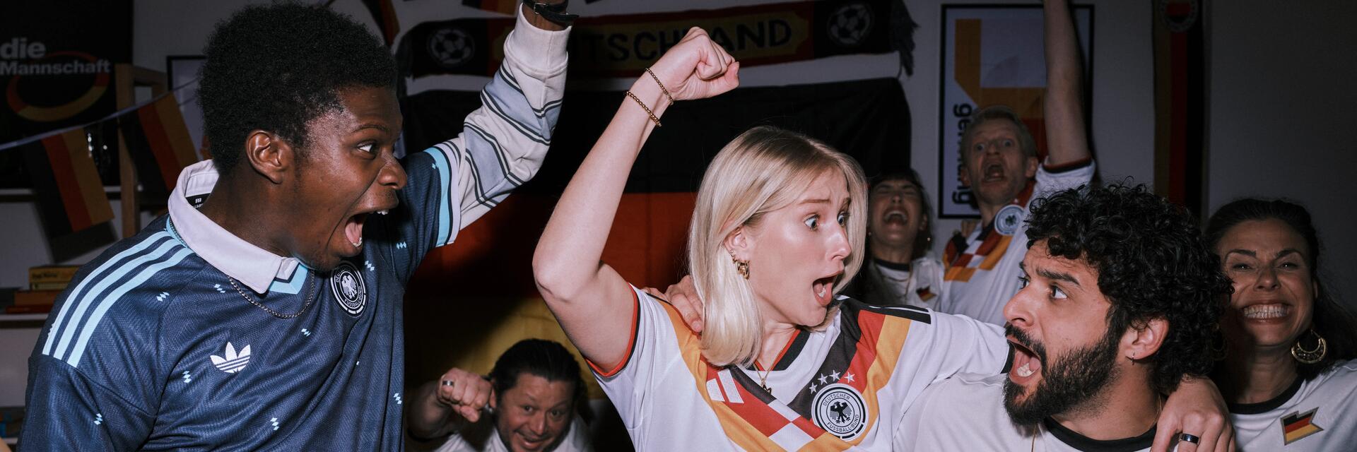 Eine Gruppe jubelnder Fußballfans feiert ausgelassen in einem mit Deutschland-Fahnen dekorierten Raum, während sie Trikots der deutschen Nationalmannschaft tragen.