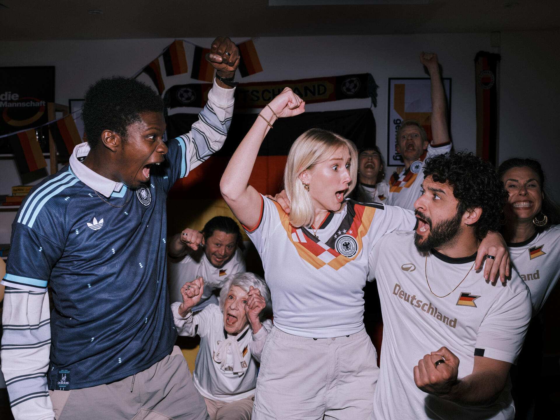 Eine Gruppe jubelnder Fußballfans feiert ausgelassen in einem mit Deutschland-Fahnen dekorierten Raum, während sie Trikots der deutschen Nationalmannschaft tragen.