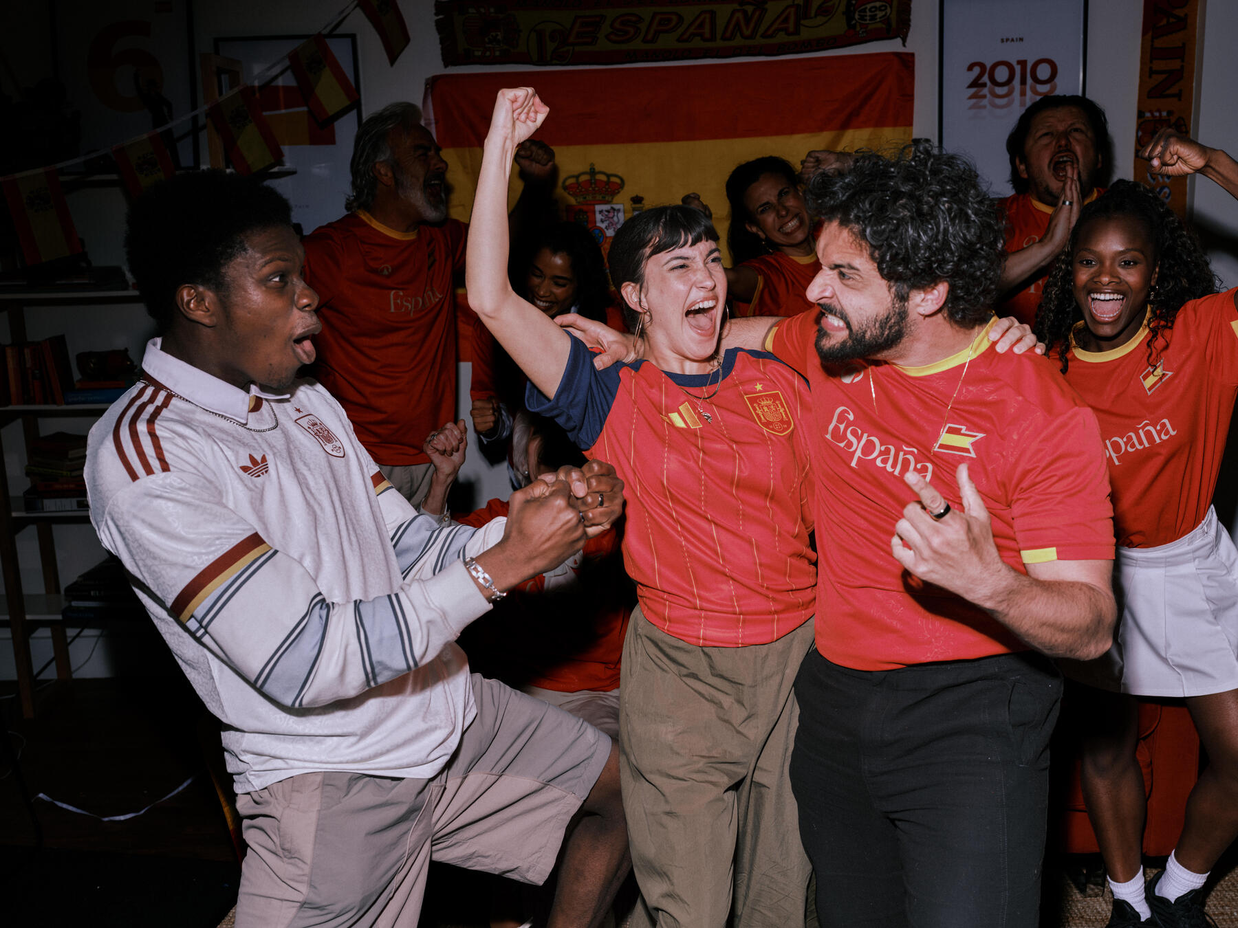 Eine Gruppe leidenschaftlicher Fans in Spanien-Trikots feiert einen emotionalen Moment in einem Raum, der mit spanischen Flaggen und Fanartikeln dekoriert ist.