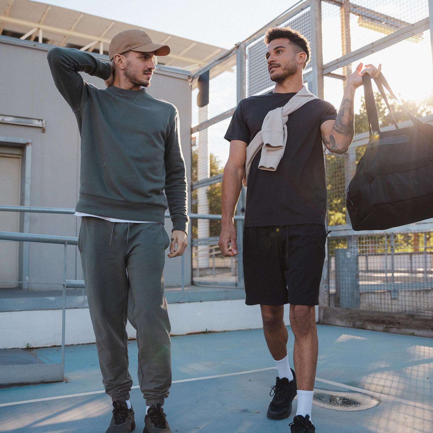 Zwei junge Männer unterhalten sich entspannt, während sie nebeneinander über einen blauen Sportplatz im Freien gehen. Der linke Mann trägt ein dunkelgraues Sweatshirt, eine passende Jogginghose, dunkle Sneaker und eine beige Basecap. Der rechte Mann trägt ein schwarzes T-Shirt, schwarze Shorts, weiße Socken und schwarze Sneaker; er hat sich einen hellen Pullover locker um die Schultern gelegt und trägt eine große schwarze Sporttasche in der Hand. Im Hintergrund sind ein graues Gebäude und ein hoher Zaun zu sehen, während die tiefstehende Sonne ein warmes Licht erzeugt.