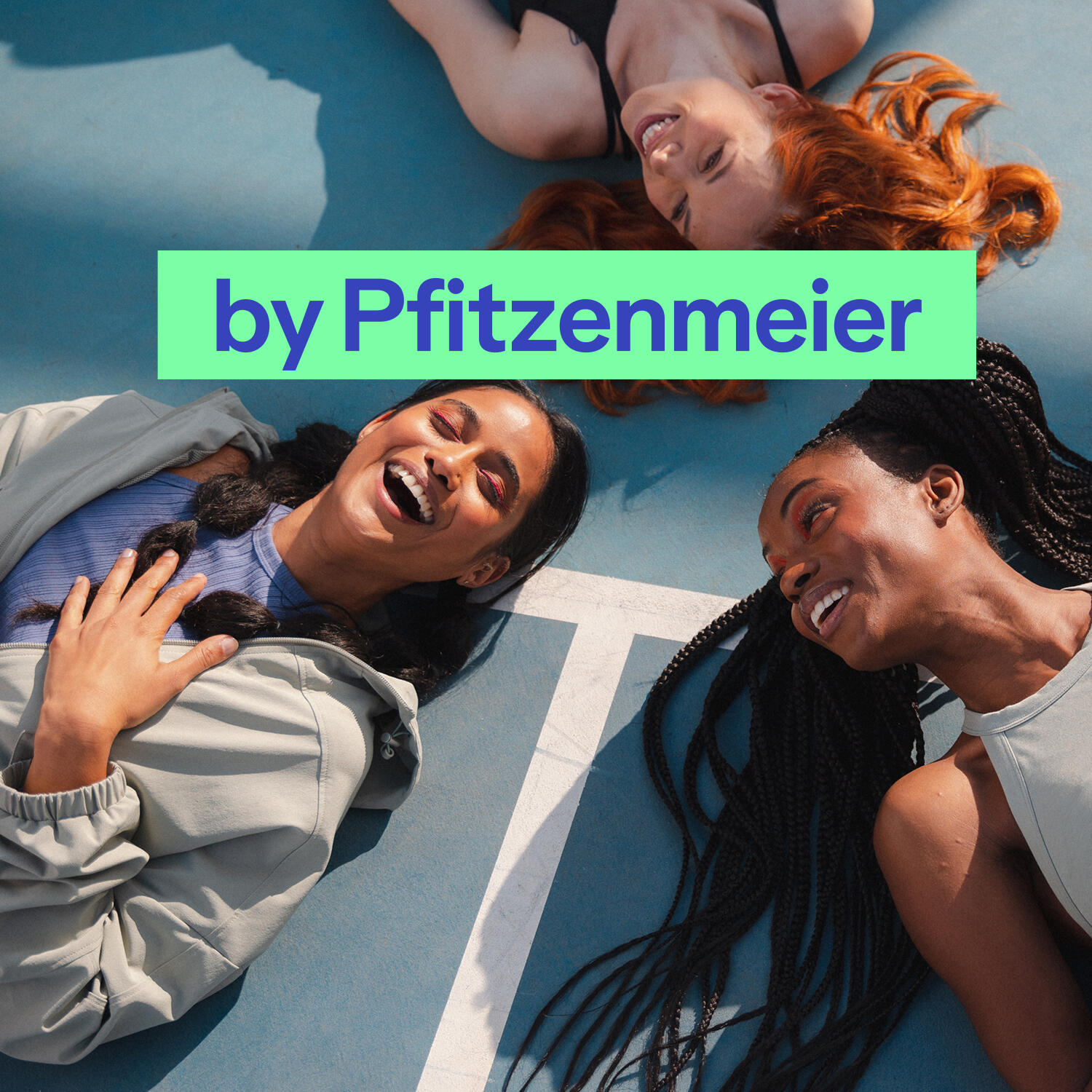 Drei junge Frauen liegen lachend in einer kreisförmigen Formation mit den Köpfen zueinander auf einem blauen Sportplatz mit weißen Markierungslinien. Die Szene ist aus der Vogelperspektive aufgenommen. Eine Frau hat langes rotes Haar, eine trägt dunkle Zöpfe und die dritte hat dunkles, zu einem Zopf geflochtenes Haar. Sie tragen sportliche Kleidung in hellen Farben und wirken ausgelassen und glücklich. Zentral über dem Bild liegt ein hellgrüner Balken mit dem dunkelblauen Text „by Pfitzenmeier“.