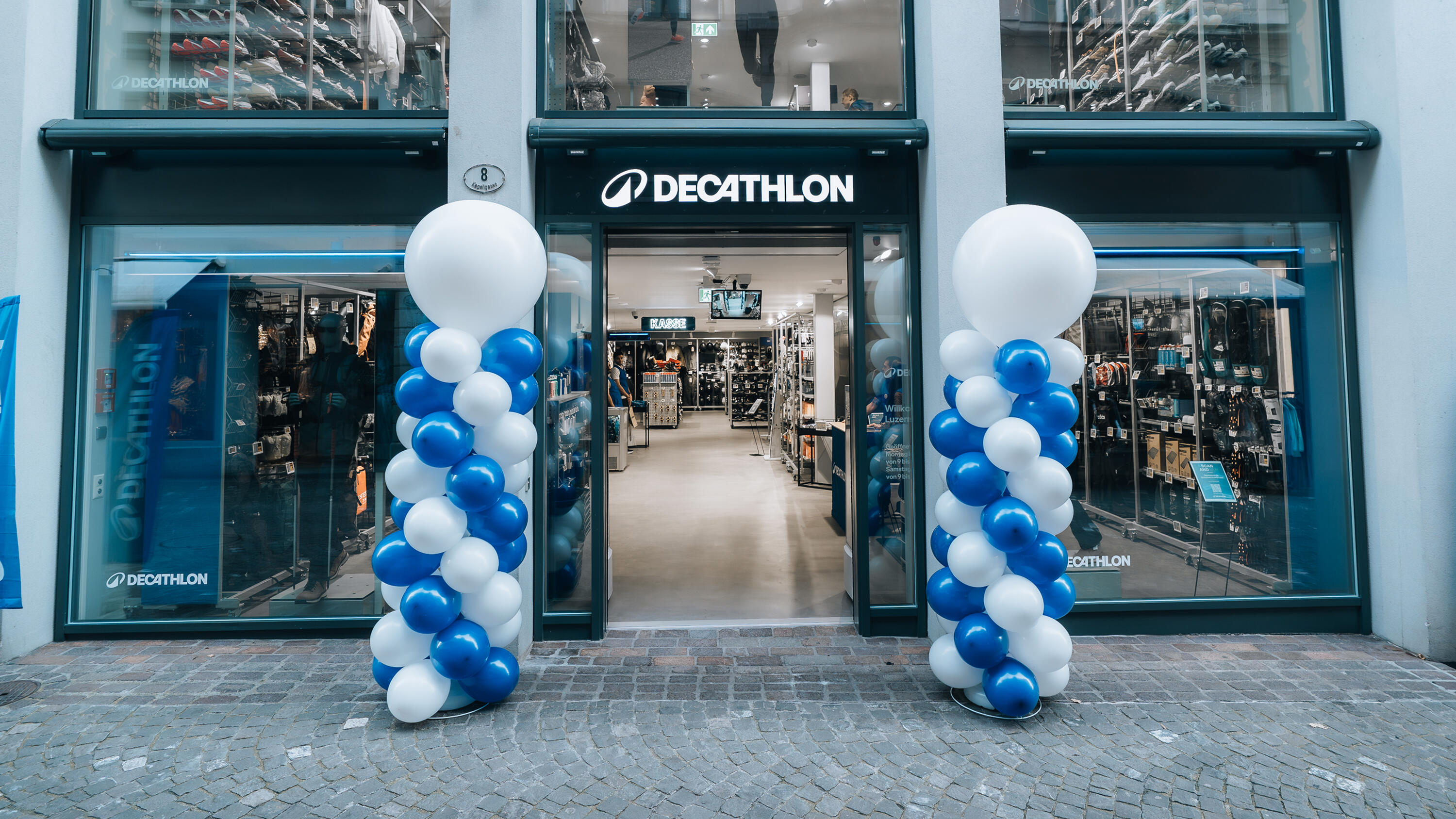 Außenansicht einer Decathlon-Filiale zur Neueröffnung. Der Eingangsbereich ist mit zwei großen Säulen aus blau-weißen Luftballons geschmückt, die jeweils von einem besonders großen weißen Ballon gekrönt werden. Über der offenen Eingangstür prangt das leuchtende weiße Decathlon-Logo auf dunklem Grund. Durch die großen Schaufenster und die Tür ist der hell erleuchtete Innenraum des Sportgeschäfts mit Regalen voller Sportartikel und Schuhe zu sehen. Der Boden vor dem Geschäft besteht aus grauem Kopfsteinpflaster.