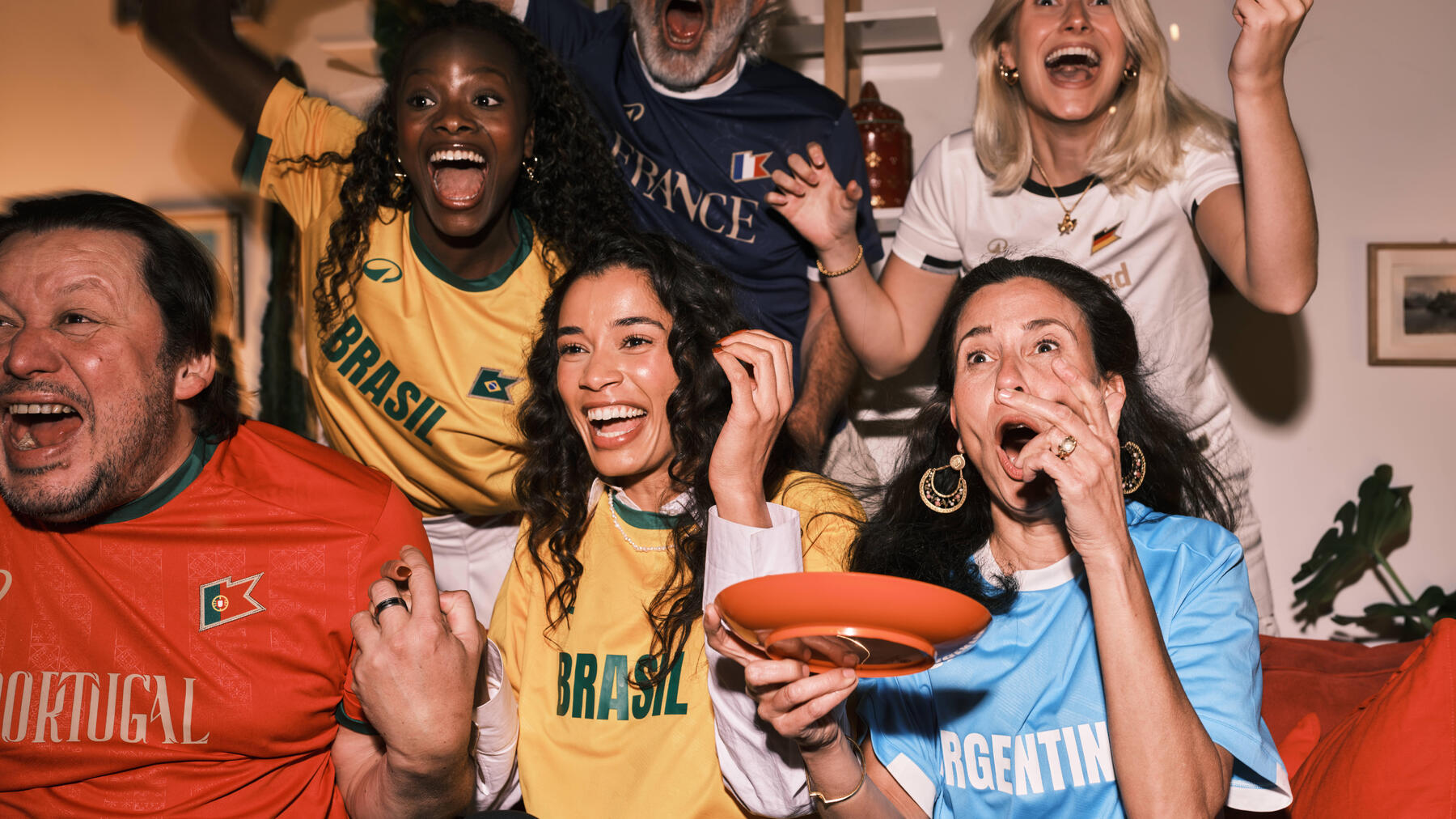 Eine Gruppe emotional mitfiebernder Fans in Trikots verschiedener Nationalmannschaften wie Brasilien, Portugal, Argentinien und Frankreich reagiert enthusiastisch auf ein Sportereignis.