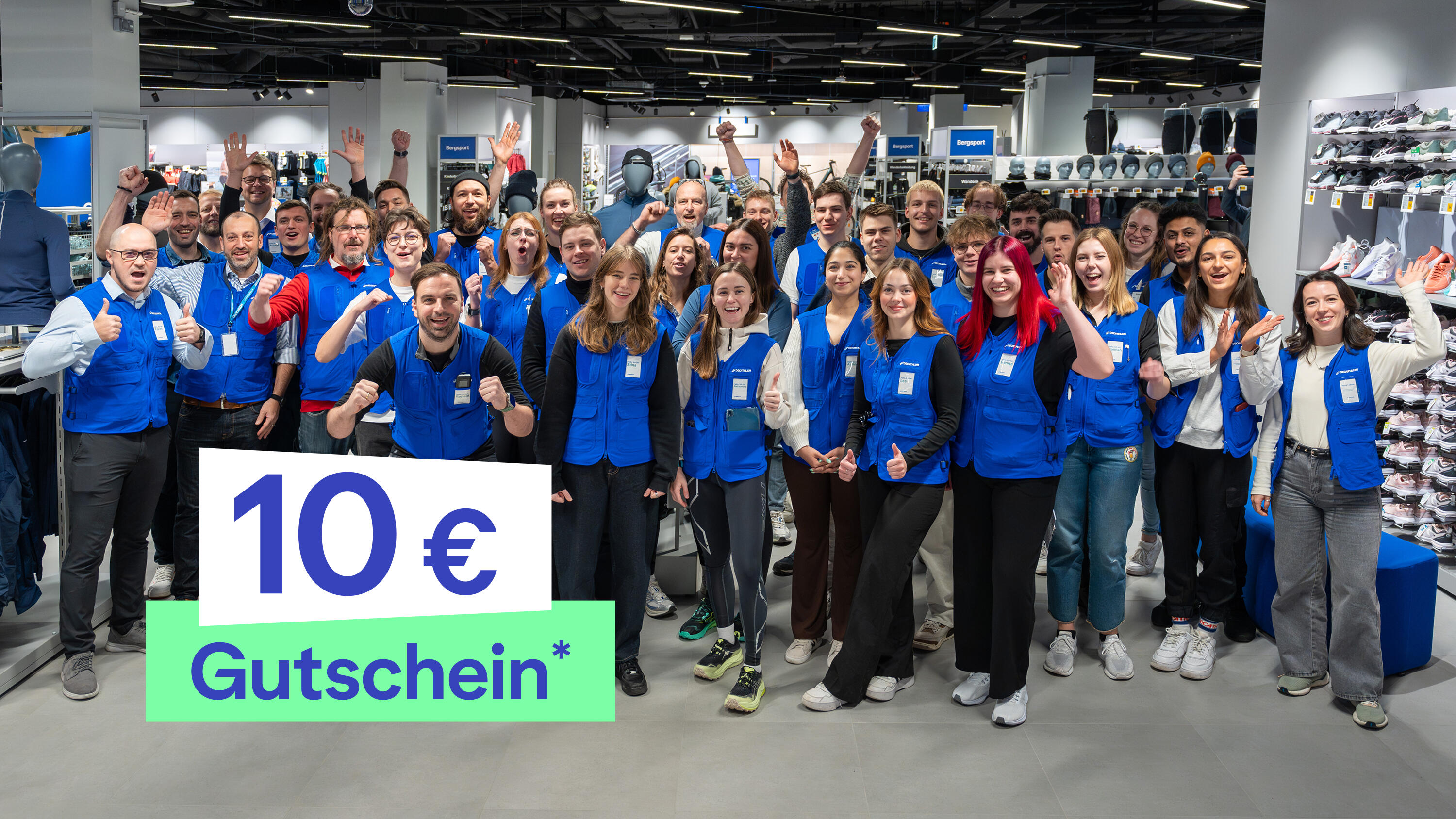 Ein großes Team von Decathlon-Mitarbeitern posiert gut gelaunt und jubelnd in einer Filiale. Viele tragen die typischen blauen Westen über ihrer Kleidung, winken oder zeigen den Daumen nach oben. Im Hintergrund sind Schuhregale und Bekleidungsständer zu sehen. Unten links im Bild ist eine Grafik mit der Aufschrift „10 € Gutschein“ eingeblendet.*