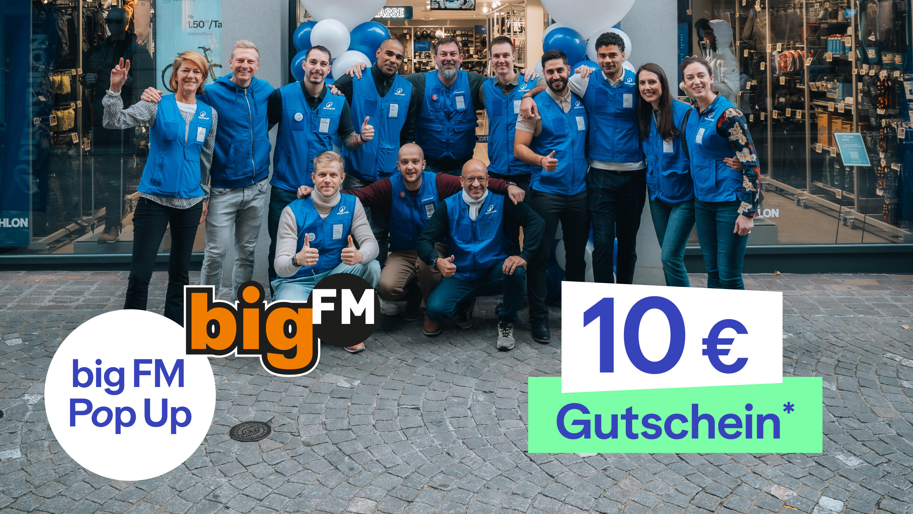 Die Gruppe: Im Zentrum steht eine Gruppe von etwa 13 gut gelaunten Menschen, die sich für ein Teamfoto aufgestellt haben – einige stehen, drei Personen knien in der vorderen Reihe. Fast alle tragen einheitliche blaue Westen über ihrer Alltagskleidung. Der Hintergrund: Die Gruppe befindet sich im Außenbereich auf grauem Kopfsteinpflaster direkt vor der Glasfront eines Geschäfts. Im Hintergrund (beim Ladeneingang) sind dekorative blau-weiße Luftballons zu sehen. Grafische Overlays (Vordergrund): Unten links: Ein weißer Kreis mit dem blauen Text „bigFM Pop Up“, direkt daneben das bekannte, orangefarbene „bigFM“-Logo. Unten rechts: Ein großes, weißes Feld mit der Aufschrift „10 €“ in blauer Schrift, gefolgt von einem hellgrünen Kasten mit dem weißen Wort „Gutschein*“.