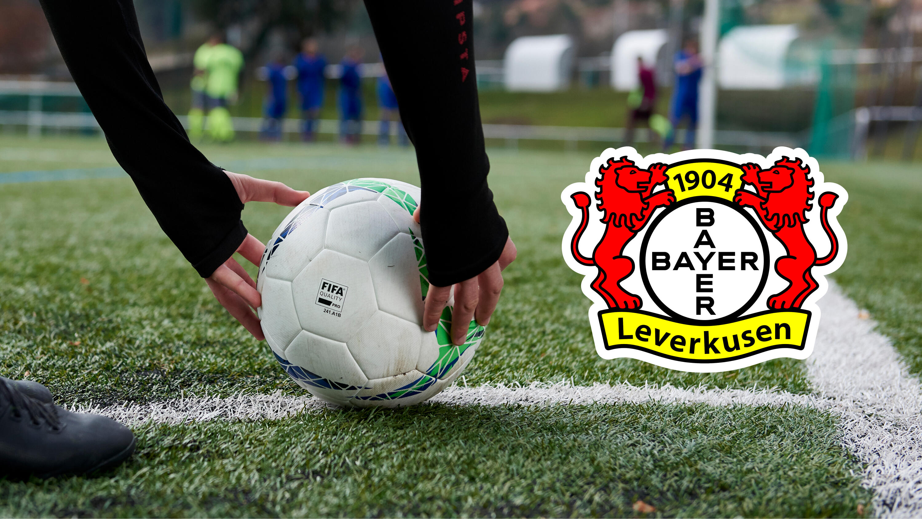Hände legen einen Fußball auf die weiße Linie eines Kunstrasenplatzes, daneben das Vereinslogo von Bayer 04 Leverkusen.