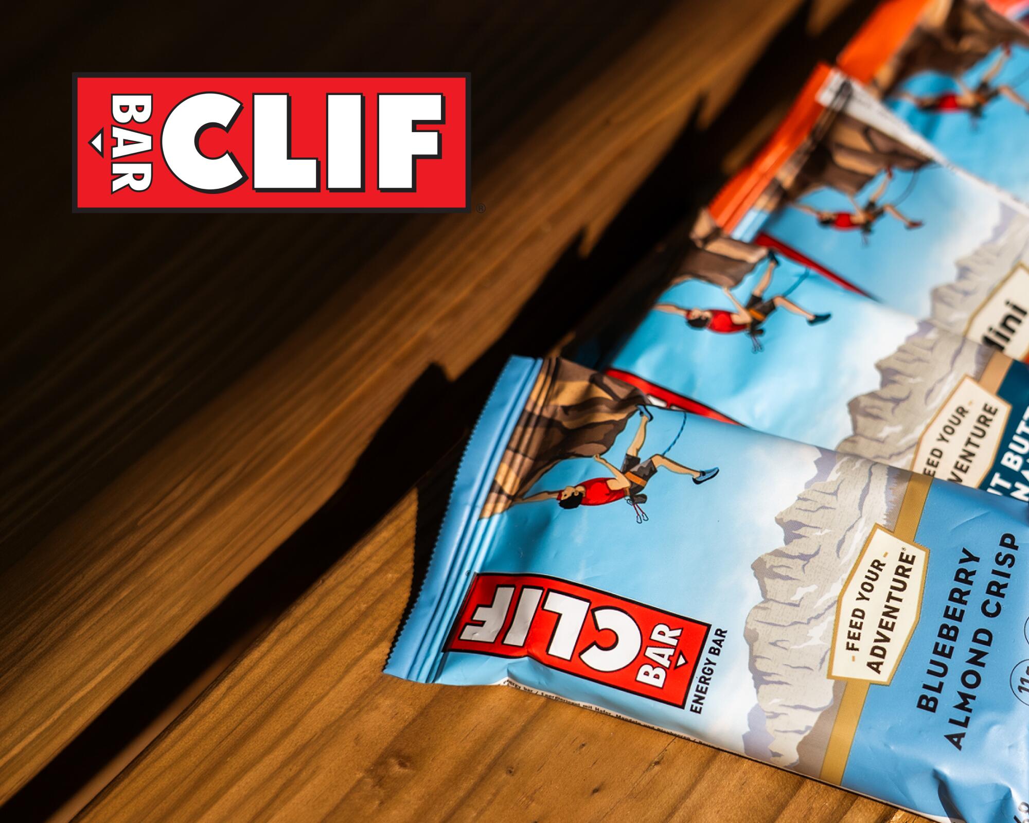 Drei CLIF Bar Müsliriegel mit Kletterer-Illustration und sichtbarer Aufschrift 'CLIF BAR' auf der Verpackung liegen auf einem Holztisch.