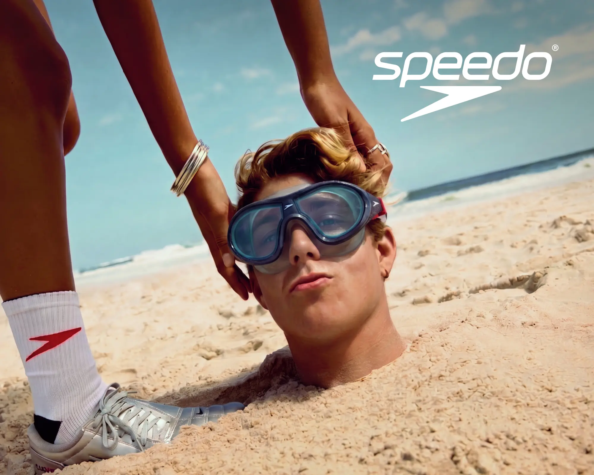 Ein junger Mann ist am Strand bis zum Hals im Sand eingegraben und trägt eine Speedo-Schwimmbrille. Neben ihm steht eine Person mit weißen Speedo-Socken. Oben rechts befindet sich das weiße Speedo-Logo.