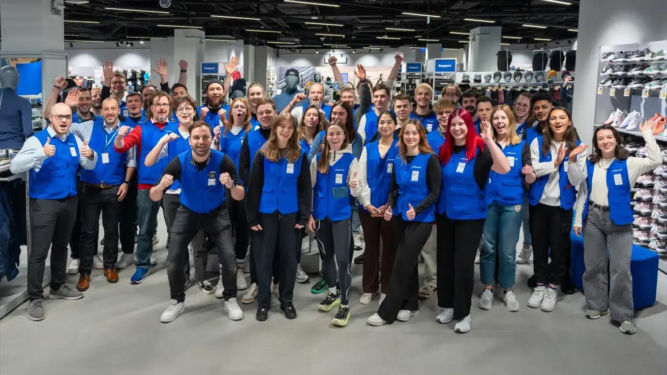 Ein Gruppenfoto eines großen Teams von Mitarbeitern, die in einem geräumigen Sportgeschäft jubeln und feiern. Die Gruppe von etwa 40 Personen, die meisten tragen blaue Firmenwesten, blickt begeistert in die Kamera. Viele haben ihre Hände oder Fäuste in die Luft gehoben, um einen feierlichen Teammoment einzufangen. Sie stehen im Gang eines hell beleuchteten Einzelhandelsgeschäfts, umgeben von Regalen mit Schuhen, Kleidung und Sportartikeln. Die Atmosphäre ist energiegeladen und positiv.