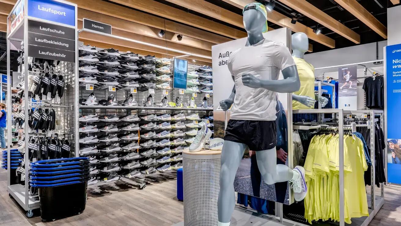 Ein Innenraumfoto der Herren-Laufabteilung eines Sportgeschäfts. Rechts im Vordergrund steht eine graue Schaufensterpuppe in dynamischer Laufpose, die ein weißes T-Shirt und schwarze Shorts trägt. Ein einzelner Laufschuh wird auf einem zylindrischen Sockel vor ihr präsentiert. Dahinter steht eine weitere graue Puppe in einem gelben T-Shirt. Eine lange Wand ist mit hohen Regalen gefüllt, die Hunderte von Laufschuhpaaren zeigen. Große Schilder darüber weisen die Bereiche aus: 'Laufsport', 'Laufschuhe', 'Laufbekleidung', 'Laufzubehör' und 'Herren'. Links sind gestapelte blaue Einkaufskörbe platziert, und rechts befinden sich Kleiderständer mit gelben Lauf-T-Shirts. Ein Bildschirm zeigt eine Laufaufnahme. Die Decke hat Holzpaneele und Strahlerbeleuchtung.