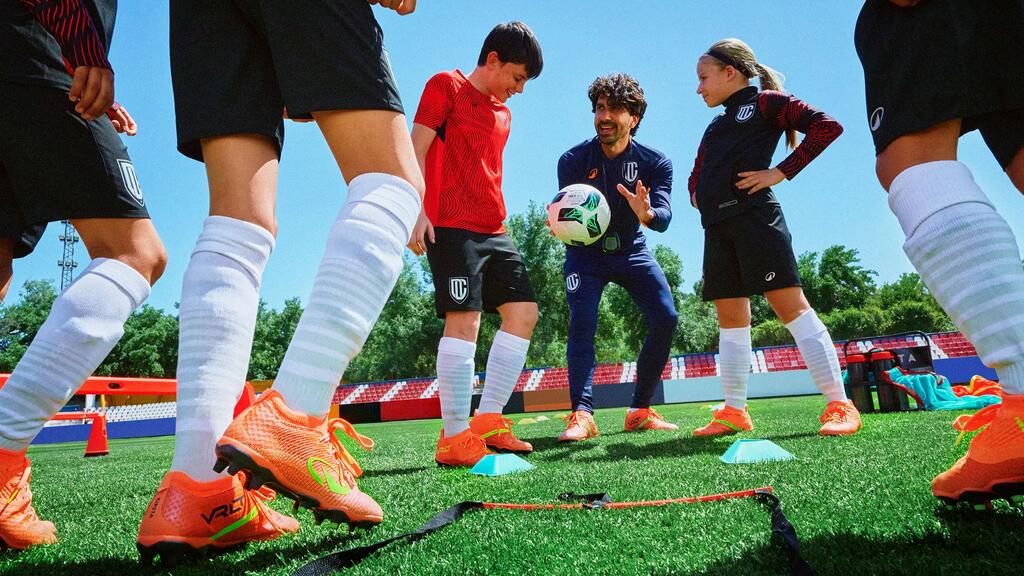 Drei begeisterte Kinder posieren Arm in Arm auf einem grünen Fußballfeld unter blauem Himmel. Sie tragen farbenfrohe Fußballtrikots in Hellgrün, Dunkelblau und Flieder mit passenden Stutzen und Sportschuhen. Mit weit geöffnetem Mund jubeln sie voller Energie direkt in die Kamera.