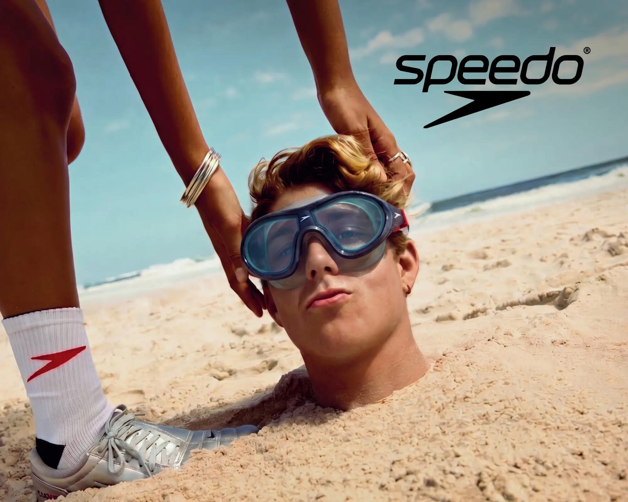 SPEEDO: Mann steckt im Sand, nur der Kopf schaut raus und er hat eine Speedo Tauchbrille auf