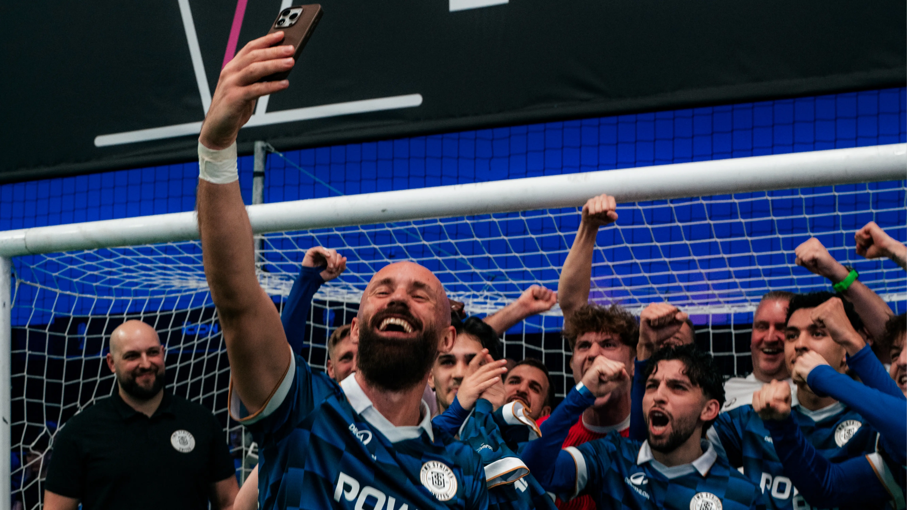 Two Stripes United Spieler jubeln vor einem Fußballtor. Der vorderste Spieler macht dabei ein Selfie mit allen.