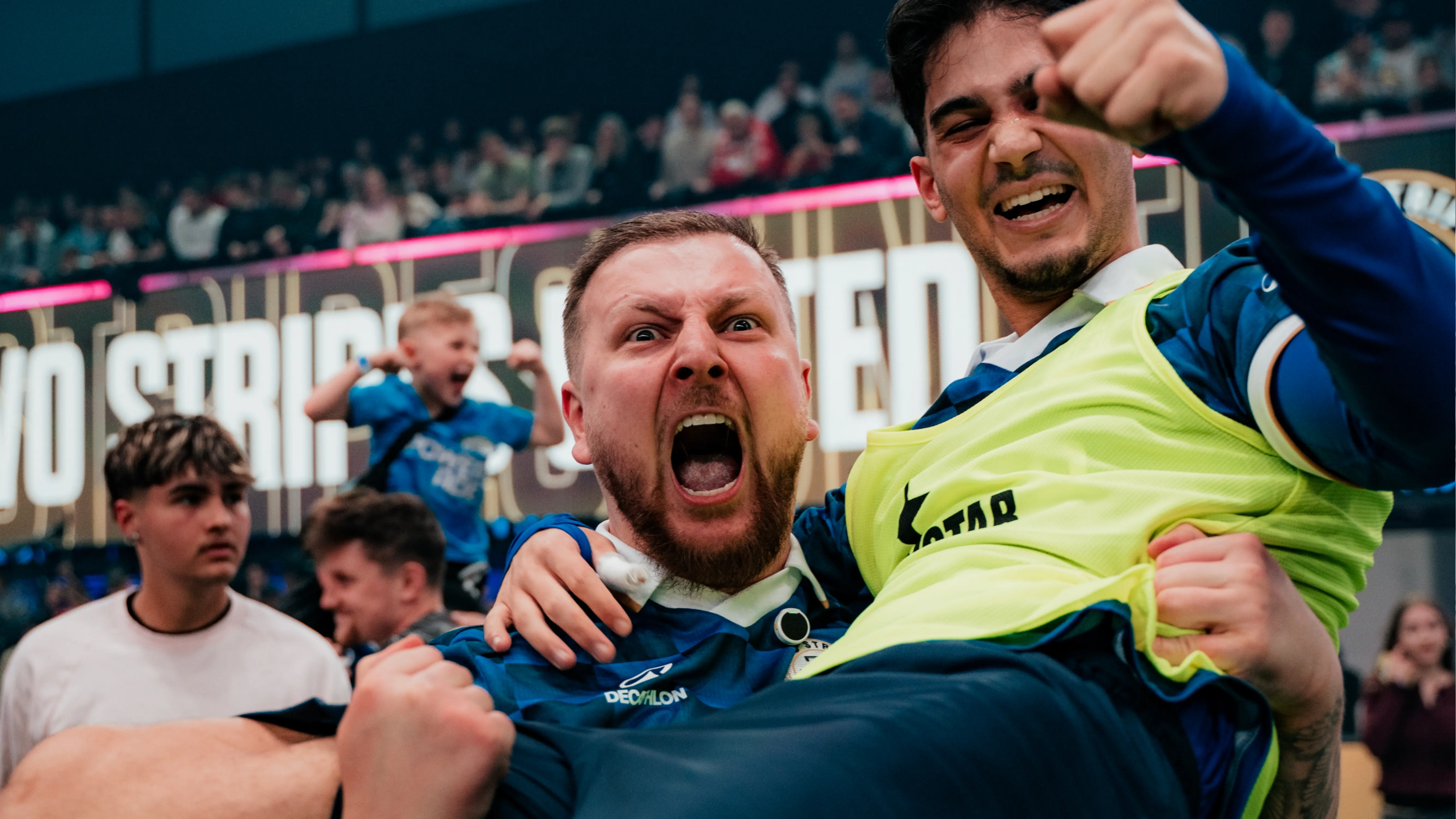 Mann in Two Stripes United trikot hält jubelnd einen Spieler von TSU in Leibchen auf dem Arm, der ebenfalls jubelt. Sie befinden sich in einer befüllten Halle.