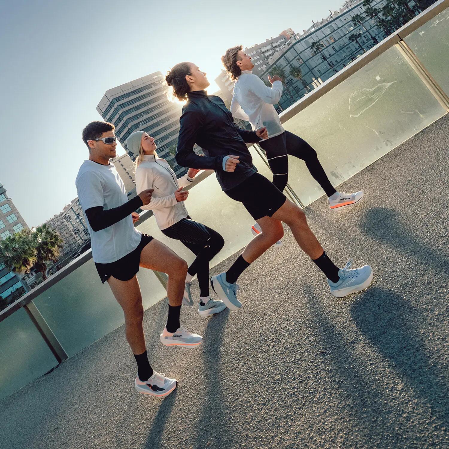 Eine Gruppe von vier Personen in Laufbekleidung joggt gemeinsam bei Sonnenschein durch eine städtische Umgebung. Sie laufen dynamisch nebeneinander über einen asphaltierten Weg mit Glasgeländer, im Hintergrund sind Gebäude, Palmen und ein blauer Himmel zu erkennen. Die Aufnahme ist aus einer leicht schrägen, niedrigen Perspektive fotografiert, was die Vorwärtsbewegung betont und lange Schatten der Läufer auf den Boden wirft. Alle tragen moderne Sportschuhe.