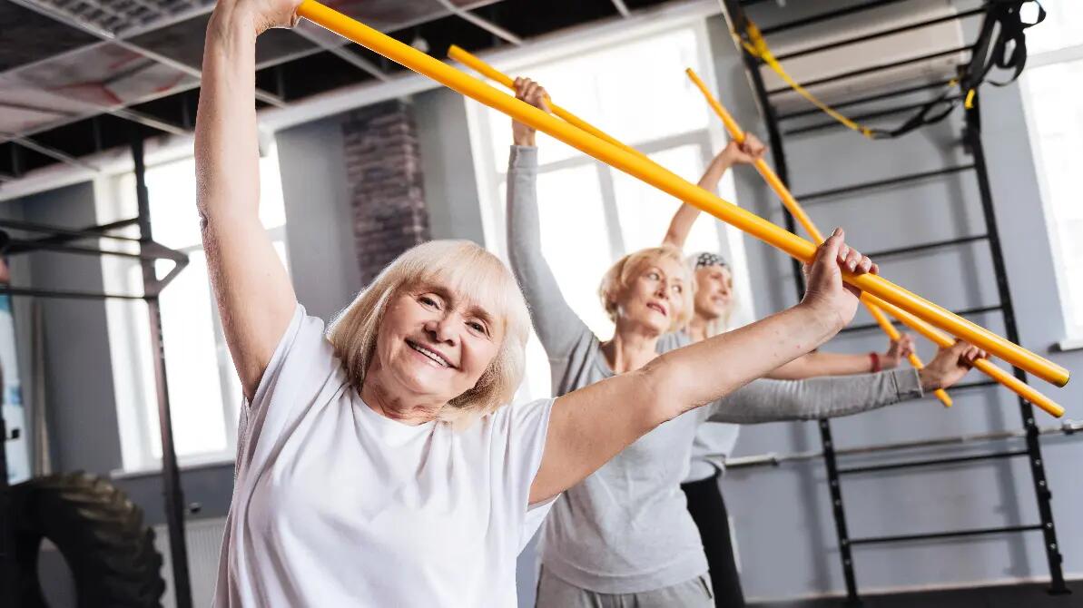 Seniorki ćwiczące z laską gimnastyczną trzymające je nad głowami