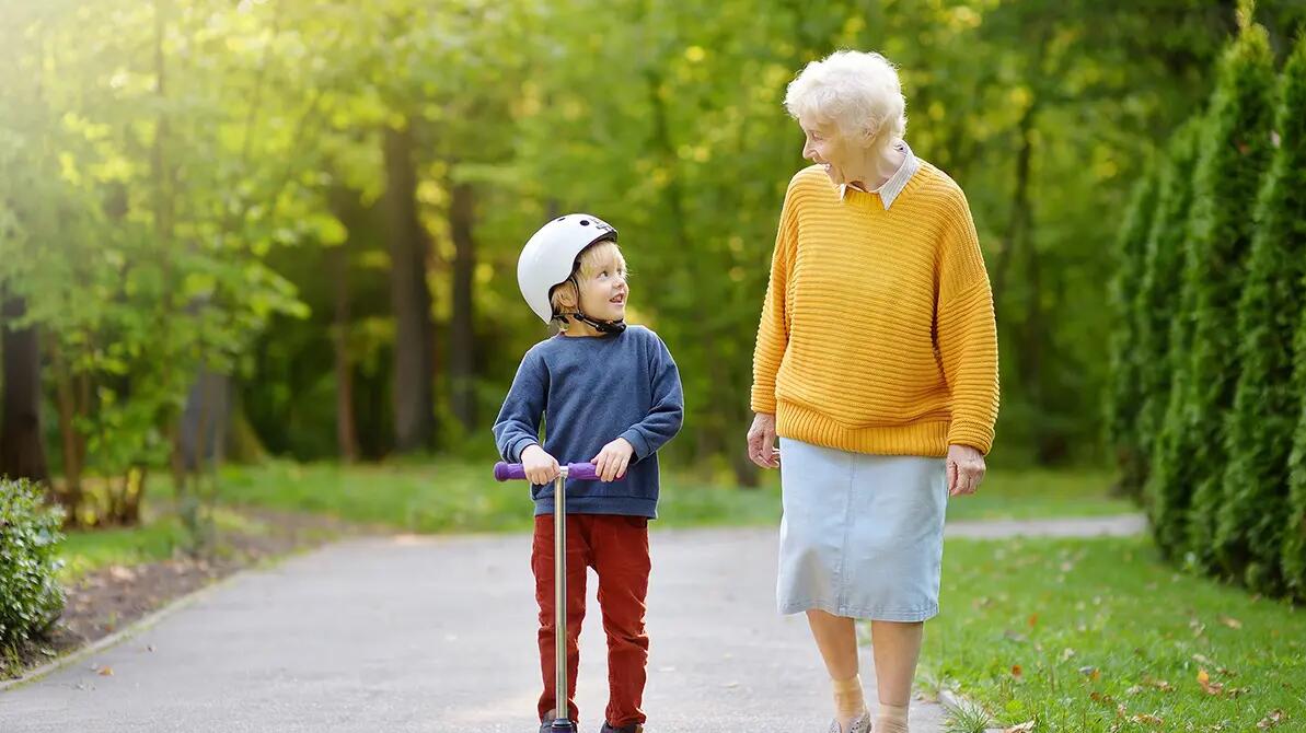Babcia idąca z wnukiem jadącym w kasku na hulajnodze w parku