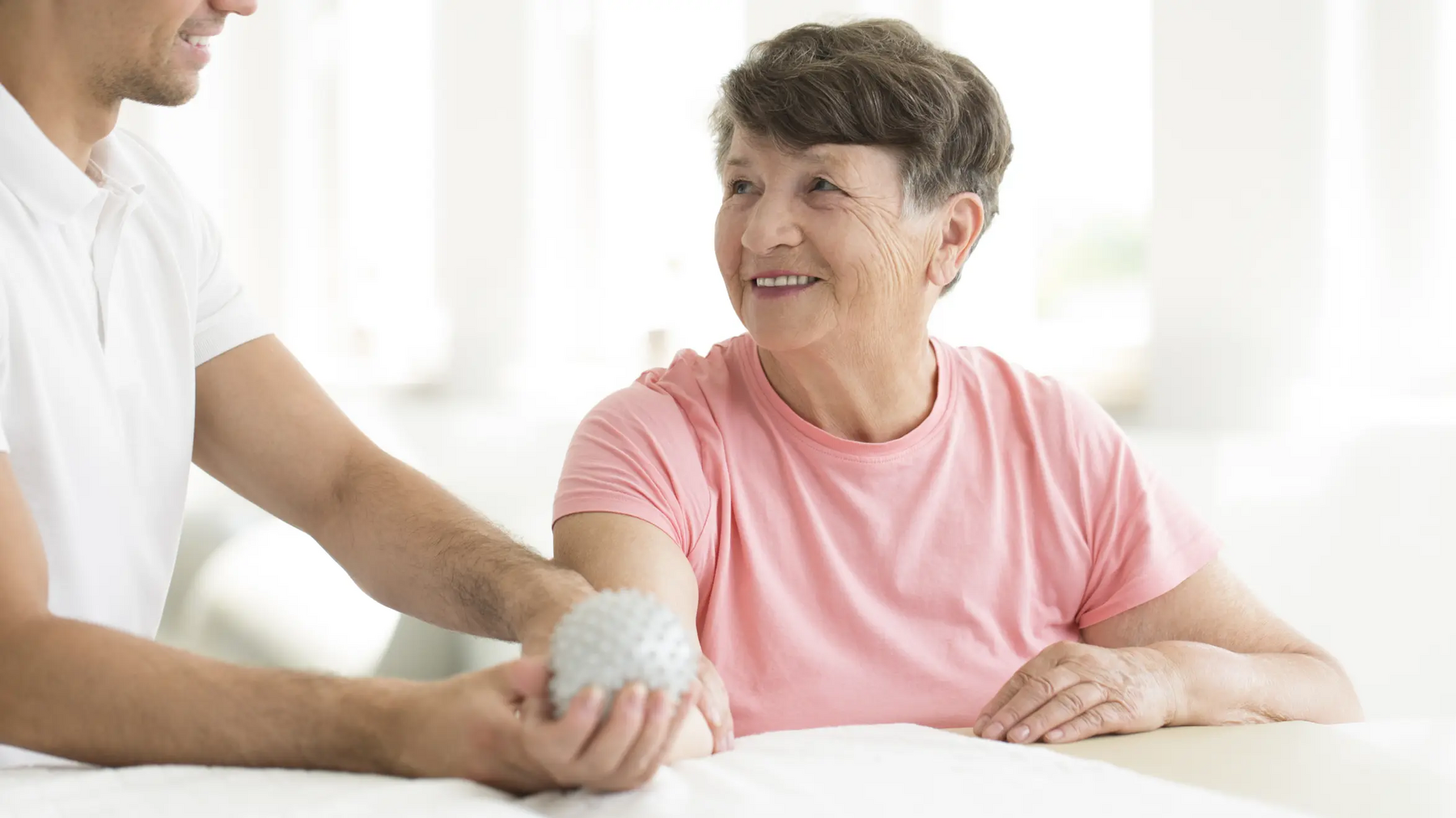 Seniorka trzymajaca w dłoni piłeczkę do masażu obok rehabiliant