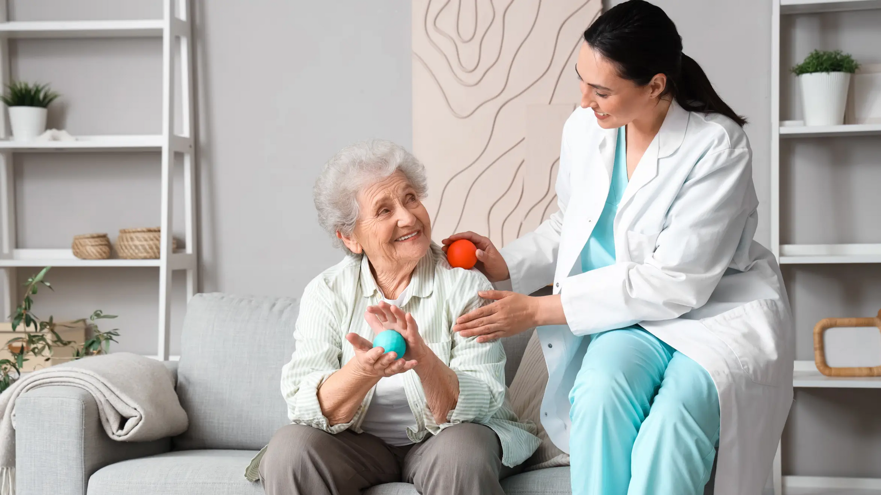Seniorka ćwiczące dłonie z piłeczką na kanapie obok siedząca rehabilitantka