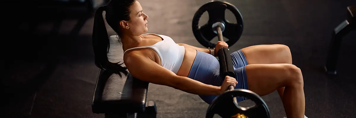 Kobieta oparta plecami o ławeczkę ze sztangą na udach ćwicząca b stance hip thrust