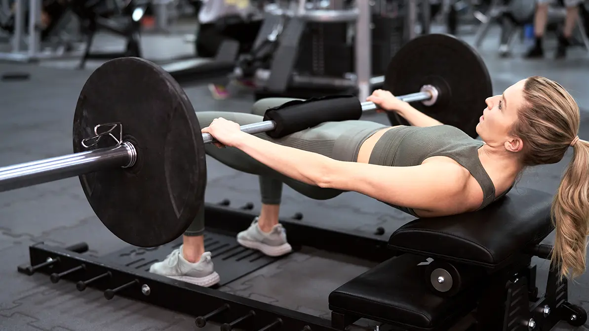 Kobieta oparta plecami o ławeczkę ze sztangą na udach ćwicząca b stance hip thrust