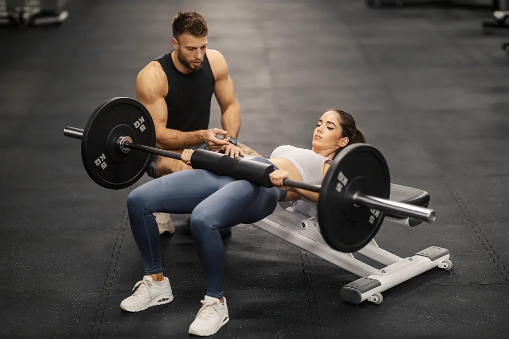 Kobieta oparta plecami o ławeczkę ze sztangą na udach ćwicząca b stance hip thrust obok trener