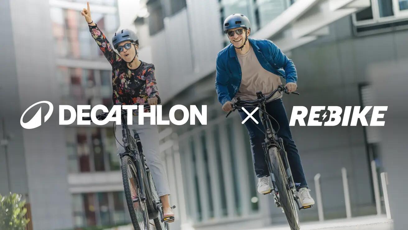 Werbebild mit dem großen weißen Schriftzug "DECATHLON X REBIKE" quer über die Bildmitte. Dahinter fahren eine Frau und ein Mann lachend auf schwarzen Fahrrädern (E-Bikes) durch eine moderne städtische Umgebung auf die Kamera zu. Beide tragen Fahrradhelme und Sonnenbrillen. Die Frau auf der linken Seite trägt ein florales Oberteil und streckt jubelnd den rechten Arm in die Höhe. Der Mann rechts trägt ein blaues Überhemd und hält beide Hände am Lenker.